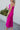 Side view of model wearing the Let's Dance Midi Dress, which features a fuchsia satin material, a cowl neckline, a sleeveless design with thin adjustable straps, a side slit, and a midi length.