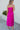 Full body back view of model wearing the Let's Dance Midi Dress, which features a fuchsia satin material, a cowl neckline, a sleeveless design with thin adjustable straps, a side slit, and a midi length.