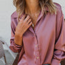 Close front view of model wearing the Always On My Mind Top, which features a mauve satin material, a collared neckline, a gold button-up front, long cuffed sleeves, a high-low design, and a flowy fit. Top is tucked into shorts.