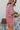 Side view of model wearing the Always On My Mind Top, which features a mauve satin material, a collared neckline, a gold button-up front, long cuffed sleeves, a high-low design, and a flowy fit. Top is tucked into shorts.