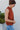 Side view of model wearing the Falling For You Top, which features a rust colored material, a rounded neck with a ruffle detail, a smocked top, a sleeveless design with thick straps, a flowy fit, and a back button closure a the neck