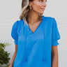 Frontal view of the Irreplaceable Top that features a royal blue material, a round neckline with a ruffle detail, a cut-out at the chest with adjustable ties, a short smocked sleeve, and a flowy fit.