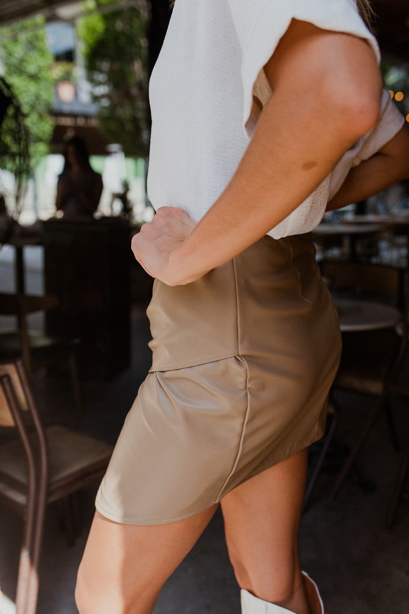 Side view of the New In Town Skirt that features a tan faux-leather material, a high-rise fit, a draped side, a surplice front, a drape side detail, a back zipper closure, and a mini length