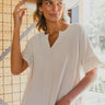 Frontal view of the At Ease Top that features an ivory colored material, a V neckline, a short cuffed sleeve, a high-low design, and a flowy fit.