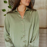 Frontal view of the On To The Next Top that features an olive green satin material, a collar neckline, a long sleeve with feather detailing at the cuff, a button-up front, and a flowy fit.