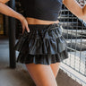 Frontal view of the One To Watch Skort that features a black faux-leather material, a thick elastic waist band with a tie detail, a tiered ruffle design, a shorts lining, and a flowy fit.