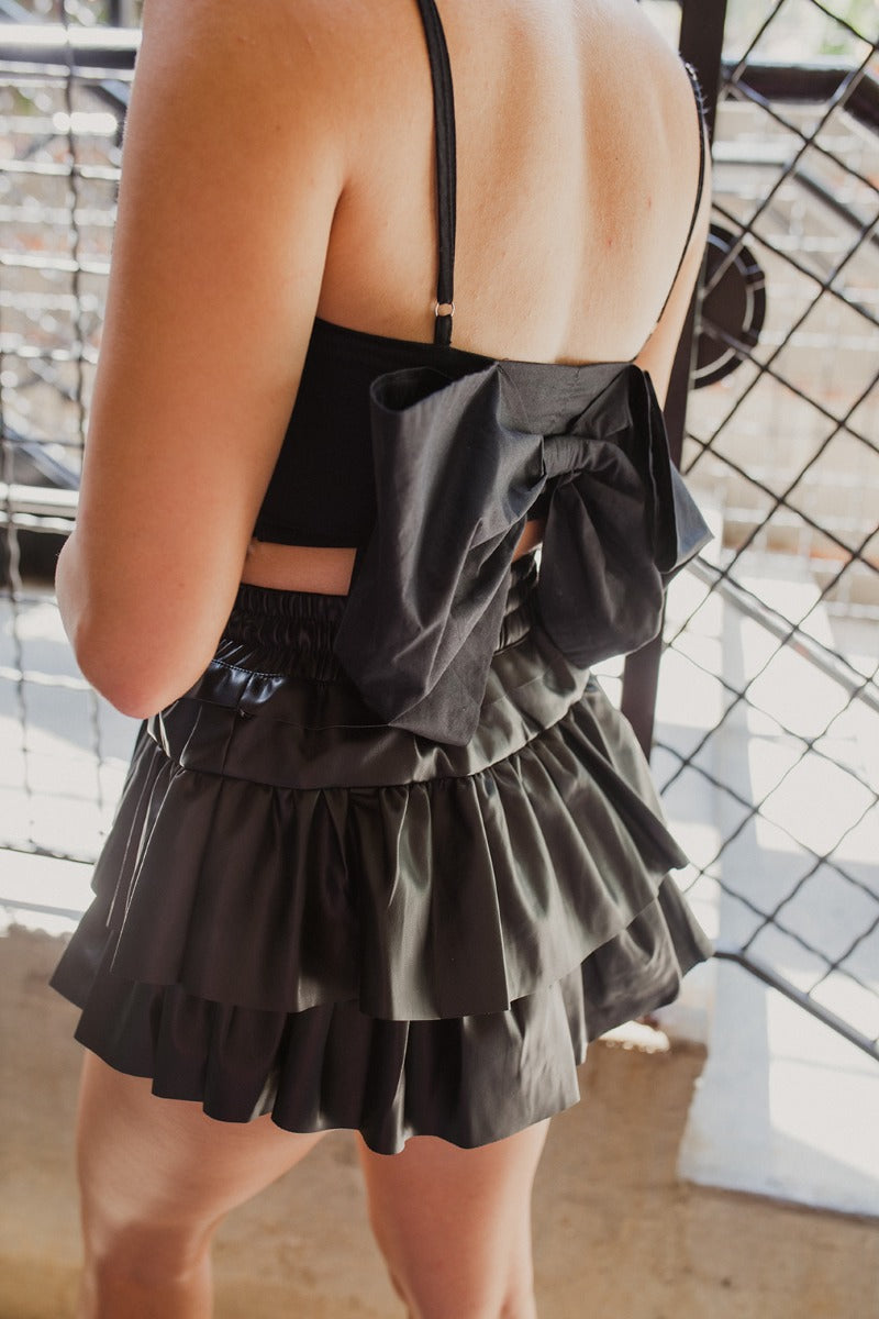 Back view of the One To Watch Skort that features a black faux-leather material, a thick elastic waist band with a tie detail, a tiered ruffle design, a shorts lining, and a flowy fit.