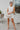 Frontal view of the Cut Out To Be Mini Dress that features a white stretchy material, a round neckline, a sleeveless design with thick straps, sewn in shoulder pads, a draped design through out, a side cut-out, and a mini length.