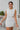 Frontal view of the Cut Out To Be Mini Dress that features a white stretchy material, a round neckline, a sleeveless design with thick straps, sewn in shoulder pads, a draped design through out, a side cut-out, and a mini length.