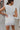 Back view of the Cut Out To Be Mini Dress that features a white stretchy material, a round neckline, a sleeveless design with thick straps, sewn in shoulder pads, a draped design through out, a side cut-out, and a mini length.
