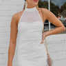 Frontal View of the Seek It Out Mini Dress that features a white colored material, a sleeveless design with a high halter neck, a mesh material at the chest, an open back, and a back zipper closure.