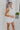 Frontal View of the Seek It Out Mini Dress that features a white colored material, a sleeveless design with a high halter neck, a mesh material at the chest, an open back, and a back zipper closure.