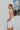 Side View of the Seek It Out Mini Dress that features a white colored material, a sleeveless design with a high halter neck, a mesh material at the chest, an open back, and a back zipper closure.