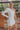 Frontal view of the Late Night Talk Dress that features white fabric, an asymmetrical neckline, a one shoulder long sleeve, a front slit, a side zipper closure, and a mini length.