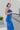 Side view of the Royalty Midi Dress that features a royal blue silk material, a sleeveless design with thin adjustable straps, a bustier design, a back zipper closure, and a midi length