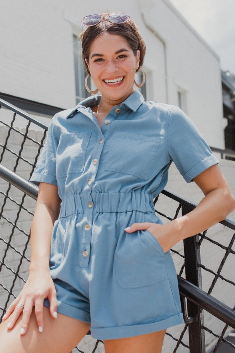 Frontal View of the Heading South Romper that features a blue colored material, a collar neckline, a short cuffed sleeve, a button-up front, two front pockets at the chest, a cinched waist, two side pockets, cuffed bottom hem, and two back pockets.