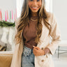 Front view of model wearing the Working On It Blazer in Beige that has a khaki colored material with a full lining, a collar and lapel, cuffed sleeves, decorative tortoise buttons, and faux front pockets. Worn over brown top.