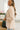 Side view of model wearing the Working On It Blazer in Beige that has a khaki colored material with a full lining, a collar and lapel, cuffed sleeves, decorative tortoise buttons, and faux front pockets. Worn over brown top.