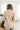 Back view of model wearing the Working On It Blazer in Beige that has a khaki colored material with a full lining, a collar and lapel, cuffed sleeves, decorative tortoise buttons, and faux front pockets. Worn over brown top.