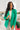 Front view of model wearing the Business As Usual Blazer in Green that has a green material with a full lining, a lapel and a collar, shoulder pads, cuffed sleeves, and faux front pockets. Worn over white top.