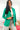 Front view of model wearing the Business As Usual Blazer in Green that has a green material with a full lining, a lapel and a collar, shoulder pads, cuffed sleeves, and faux front pockets. Worn over white top.