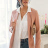 Front view of model wearing the Business As Usual Blazer in Blush, that has  a dusty pink material with a full lining, a lapel and a collar, shoulder pads, cuffed sleeves, and faux front pockets. Worn over white blouse.