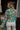 Back view of model wearing the Having A Good Day Top that has turquoise plisse fabric with a multicolor floral pattern, a ruffled hem, a cropped waist, a button-up front, a collar, and long sleeves. Worn with one button buttoned.
