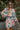 Front view of model wearing the Look On The Bright Side Dress that has a pink, green, blue, yellow and cream flower pattern, a mini-length hem, an elastic waist with a tie, a surplice neck, and long balloon sleeves.