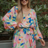 Front view of model wearing the Look On The Bright Side Dress that has a pink, green, blue, yellow and cream flower pattern, a mini-length hem, an elastic waist with a tie, a surplice neck, and long balloon sleeves.