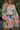 Close front view of model wearing the Look On The Bright Side Dress that has a pink, green, blue, yellow and cream flower pattern, a mini-length hem, an elastic waist with a tie, a surplice neck, and long balloon sleeves.