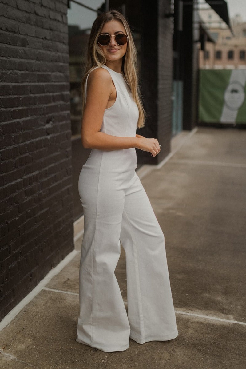 Side view of model wearing the Ready To Go Denim Jumpsuit that has white denim fabric, a round neckline, a thick waistband, a back zipper and hook closure, a sleeveless body, and wide flared legs