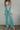 Front view of model wearing the Let's Chat Pants in Teal that have light turquoise fabric, exposed seams down the front legs, a v-shaped waistline, a monochromatic back zipper, and flared legs