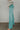 Side view of model wearing the Let's Chat Pants in Teal that have light turquoise fabric, exposed seams down the front legs, a v-shaped waistline, a monochromatic back zipper, and flared legs