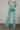 Back view of model wearing the Let's Chat Pants in Teal that have light turquoise fabric, exposed seams down the front legs, a v-shaped waistline, a monochromatic back zipper, and flared legs