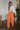 Full body front view of model wearing the Pop Of Color Pants that feature orange fabric, side pockets, a front zipper with a hook closure, and wide legs with pleated details.