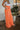 Side view of model wearing the Pop Of Color Pants that feature orange fabric, side pockets, a front zipper with a hook closure, and wide legs with pleated details.