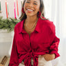 Front view of model wearing the Bristol Oversized Satin Top in Red that has red satin fabric, a button-up front with a collared neckline, one chest pocket, long sleeves with buttoned cuffs, and an oversized fit Shown tied up.