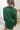 Back view of model wearing the Fancy Me Satin Top in Green which features emerald green satin fabric, a button-up front with a collared neckline, one chest pocket, and long sleeves with buttoned cuffs.