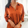 Front view of model wearing the Fancy Me Satin Top in Rust which features rust satin fabric, a button-up front with a collared neckline, one chest pocket, and long raglan sleeves with buttoned cuffs.