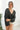Front view of model wearing the Checked Out Cropped Satin Top that has black satin fabric with a monochromatic checker print, a surplice neck, a cropped waist, and long batwing sleeves.