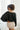 Back view of model wearing the Checked Out Cropped Satin Top that has black satin fabric with a monochromatic checker print, a surplice neck, a cropped waist, and long batwing sleeves.