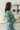 Side view of model wearing the Spot On Satin Top in Jade that has light teal satin fabric with a monochromatic spot print, a button-up front with a collar and lapels, and long sleeves with buttoned cuffs