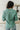 Back view of model wearing the Spot On Satin Top in Jade that has light teal satin fabric with a monochromatic spot print, a button-up front with a collar and lapels, and long sleeves with buttoned cuffs