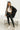 Full body Front view of model wearing the Aria Oversized Satin Top in Black that has black satin fabric, a button-up front with a collar, one pocket and long sleeves. Worn unbuttoned over white top