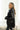 Side view of model wearing the Aria Oversized Satin Top in Black that has black satin fabric, a button-up front with a collar, one pocket and long sleeves.
