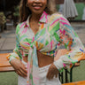 Front view of model wearing the Resort Ready Crop Top that features a pink, green, blue, yellow and cream flower pattern, a cropped tie waist, a collared neckline, and long balloon sleeves with buttoned cuffs