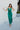 Full body front view of model wearing the Life Of The Party Maxi Dress which has an emerald green satin material, a deep open neckline, thin adjustable straps, a side drape detail with a slit, an open back, and a back zipper closure.