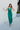 Full body front view of model wearing the Life Of The Party Maxi Dress which has an emerald green satin material, a deep open neckline, thin adjustable straps, a side drape detail with a slit, an open back, and a back zipper closure.