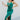 Full body side view of model wearing the Life Of The Party Maxi Dress which has an emerald green satin material, a deep open neckline, thin adjustable straps, a side drape detail with a slit, an open back, and a back zipper closure.