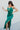 Full body side view of model wearing the Life Of The Party Maxi Dress which has an emerald green satin material, a deep open neckline, thin adjustable straps, a side drape detail with a slit, an open back, and a back zipper closure.